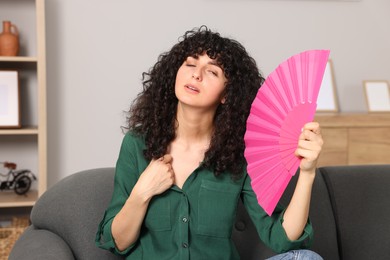 Photo of Young woman waving pink hand fan to cool herself on sofa at home