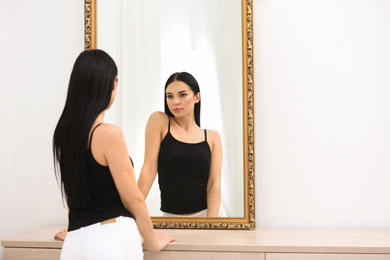 Beautiful young woman looking at herself in mirror indoors