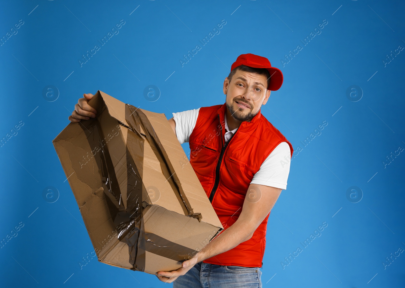 Photo of Emotional courier with damaged cardboard box on blue background. Poor quality delivery service