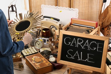 Photo of Woman with beautiful mirror at table. Sign Garage Sale written on blackboard near many different stuff indoors
