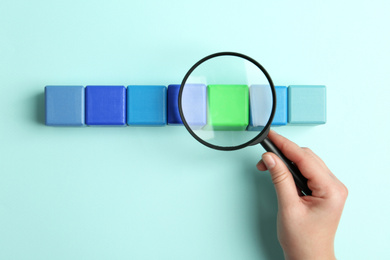 Woman holding magnifying glass above colorful cubes on light blue background, top view. Search concept
