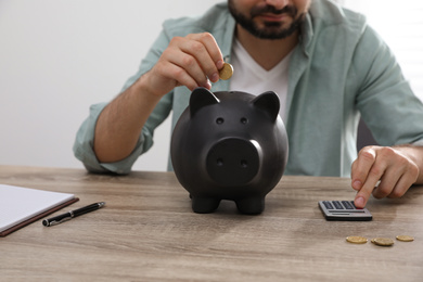 Photo of Man with calculator putting coin into piggy bank at wooden table, closeup