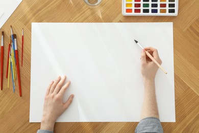 Photo of Man painting with watercolor on blank paper at wooden table, top view