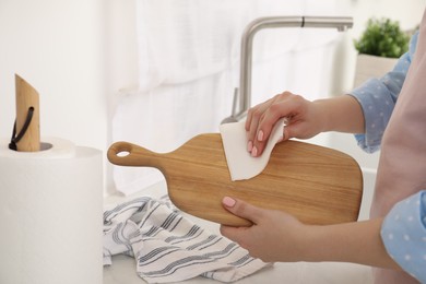 Photo of Woman wiping wooden cutting board with paper napkin in kitchen, closeup
