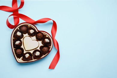 Heart shaped box with delicious chocolate candies and ribbon on light blue background, flat lay. Space for text