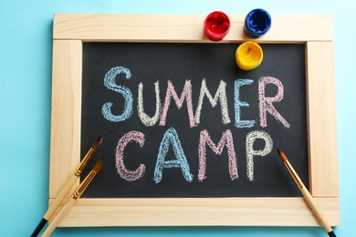 Photo of Text "SUMMER CAMP" on chalkboard, brushes and paints, top view