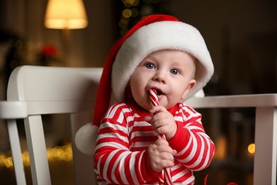 Cute baby in Santa hat and bright Christmas pajamas eating candy cane at home