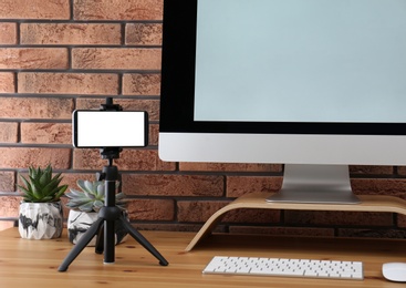 Tripod with smartphone near computer on wooden table indoors. Mockup for design