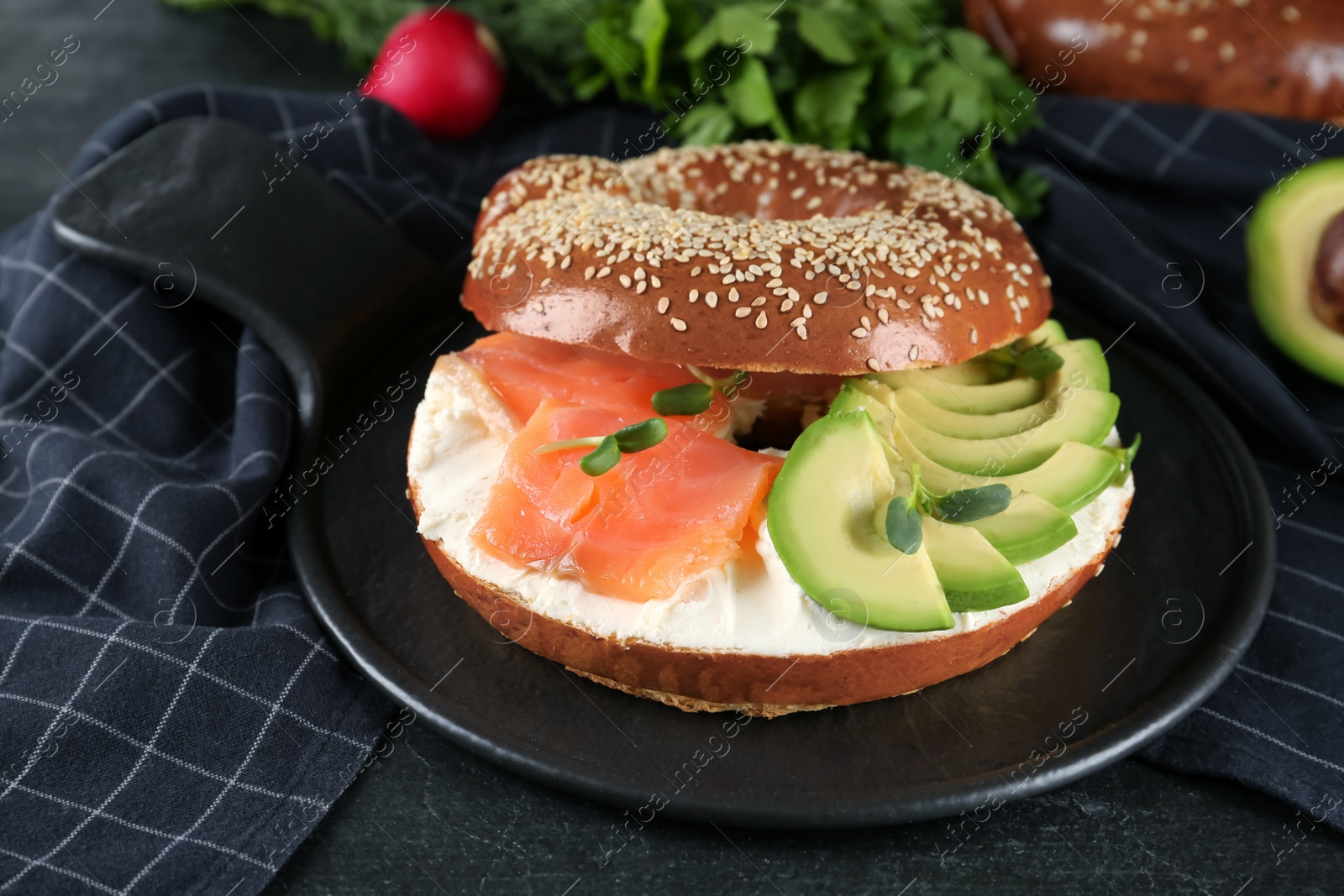 Photo of Delicious bagel with cream cheese, salmon, avocado and microgreens on black table
