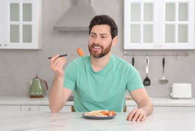 Photo of Happy man holding fork with sausage and pasta at table in kitchen