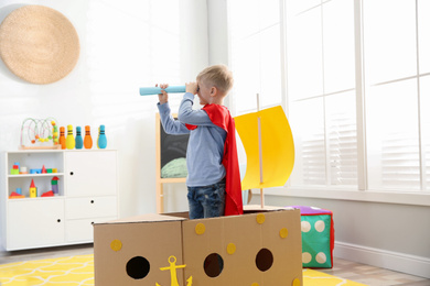 Photo of Little child in red cape playing with ship made of cardboard box at home