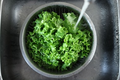 Photo of Pouring tap water into colander with lettuce in sink, top view