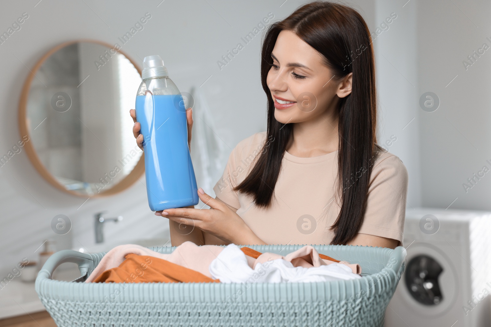 Photo of Woman holding fabric softener near basket with dirty clothes in bathroom