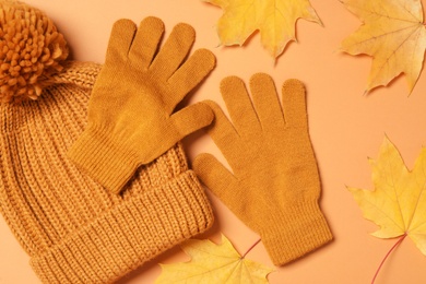 Stylish woolen gloves, hat and dry leaves on pale orange background, flat lay