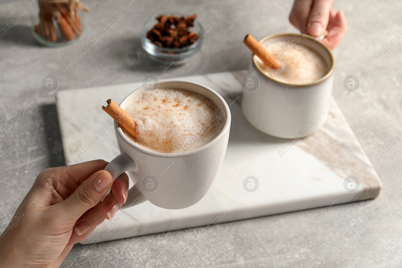 Photo of Women holding glass cups of delicious eggnog with cinnamon and anise at light grey table, closeup