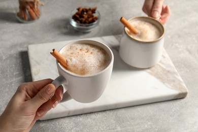 Photo of Women holding glass cups of delicious eggnog with cinnamon and anise at light grey table, closeup