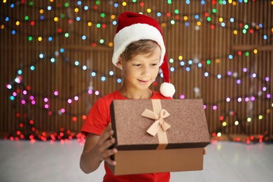 Photo of Cute little child in Santa hat opening Christmas gift box on blurred lights background
