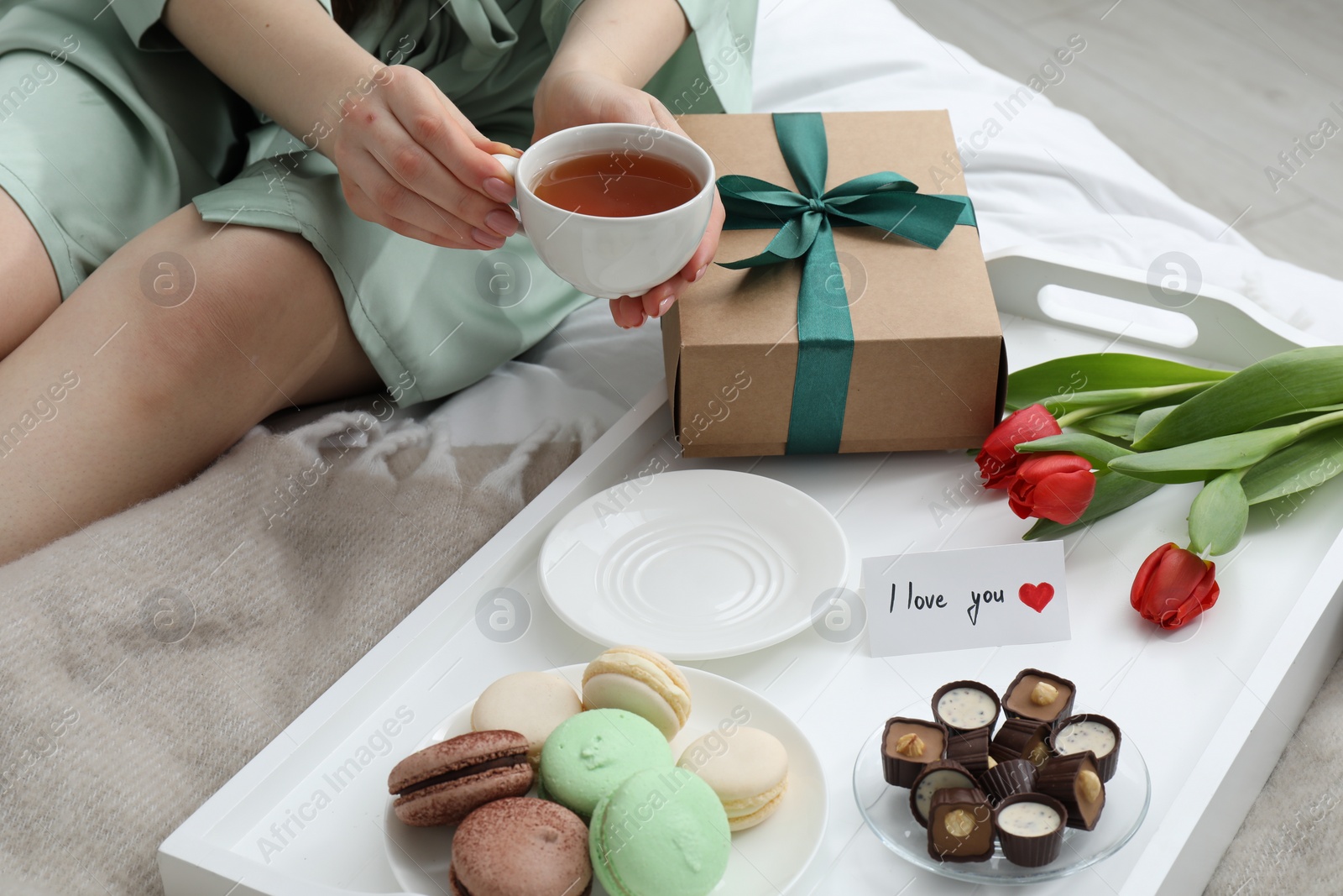 Photo of Tasty breakfast served in bed. Woman with tea, desserts, gift box, flowers and I Love You card at home, closeup