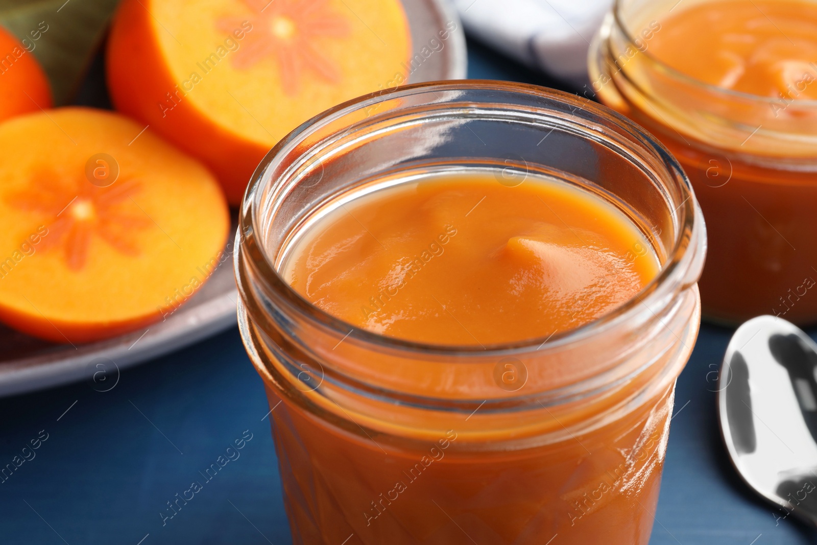 Photo of Delicious persimmon jam in glass jar and fresh fruits on blue table, closeup