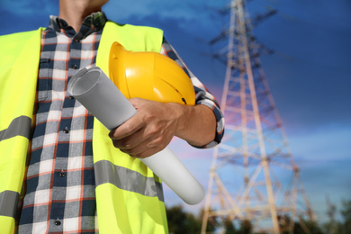Photo of Professional electrician with drafting and helmet near high voltage tower, closeup