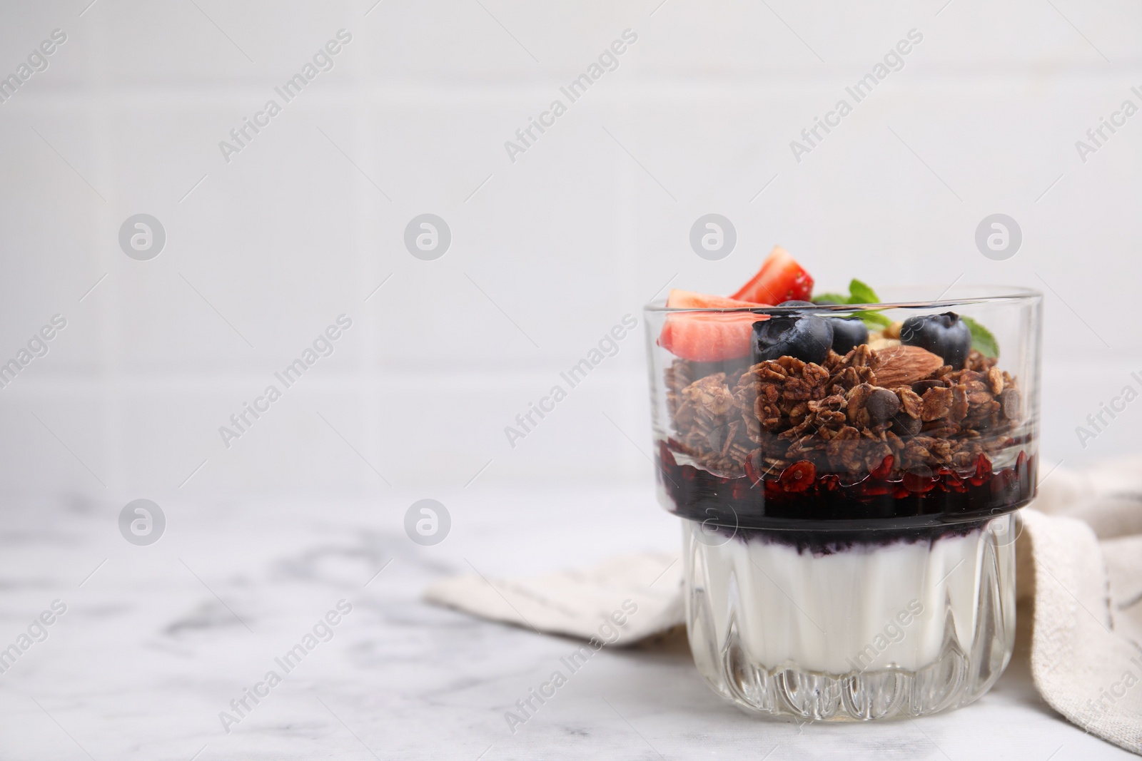 Photo of Tasty granola with berries, jam and yogurt in glass on white marble table, space for text