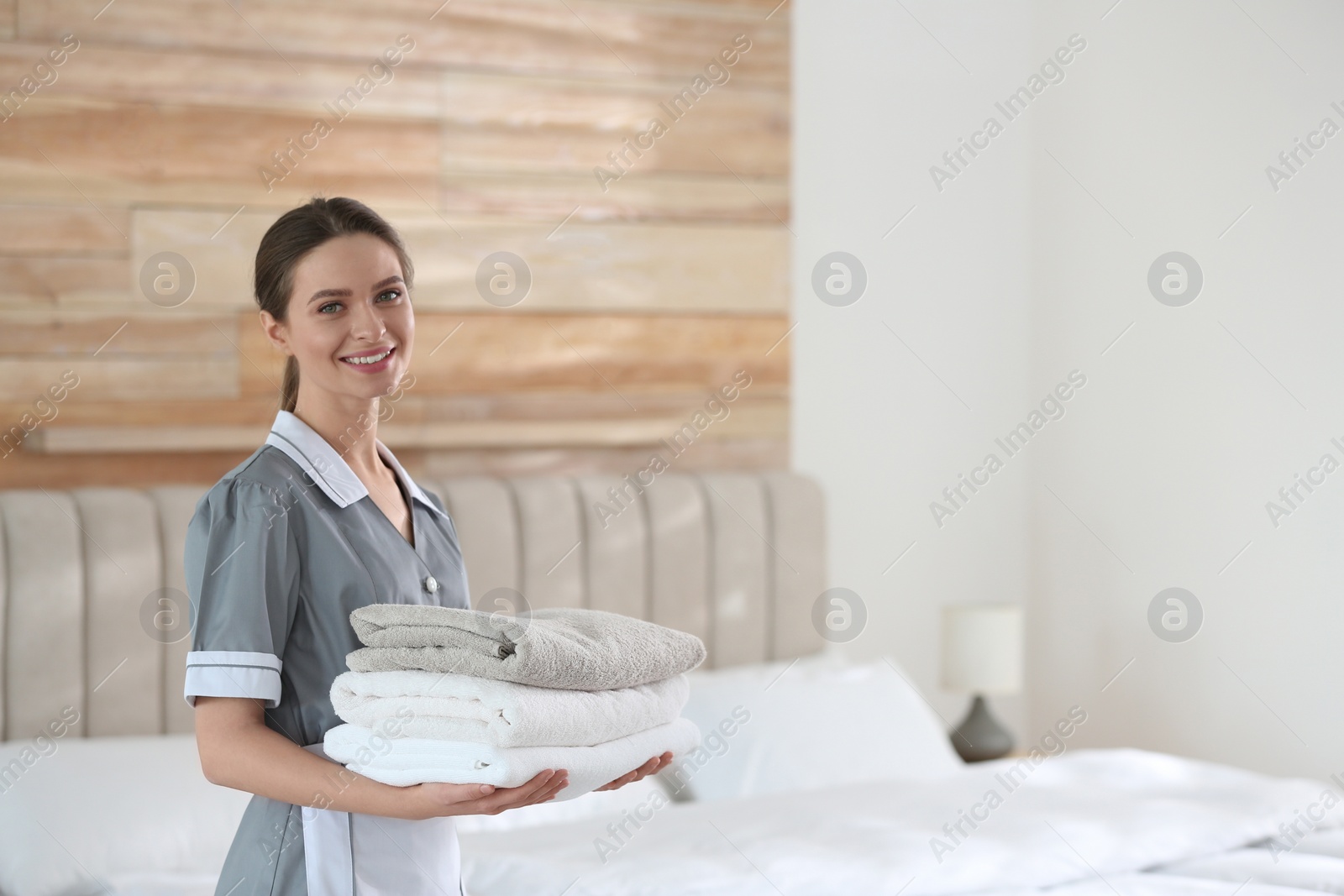 Photo of Young chambermaid holding stack of fresh towels in bedroom. Space for text