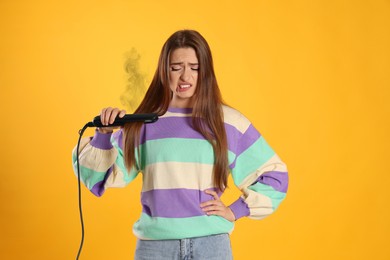 Upset young woman with flattening iron on yellow background. Hair damage