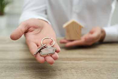 Photo of Real estate agent holding house key with trinket at wooden table, closeup