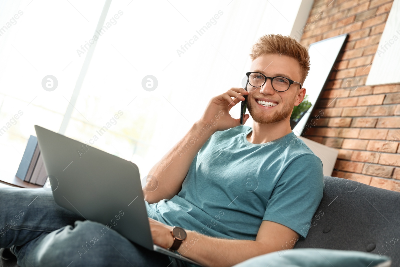 Photo of Young man talking on phone while using laptop in living room