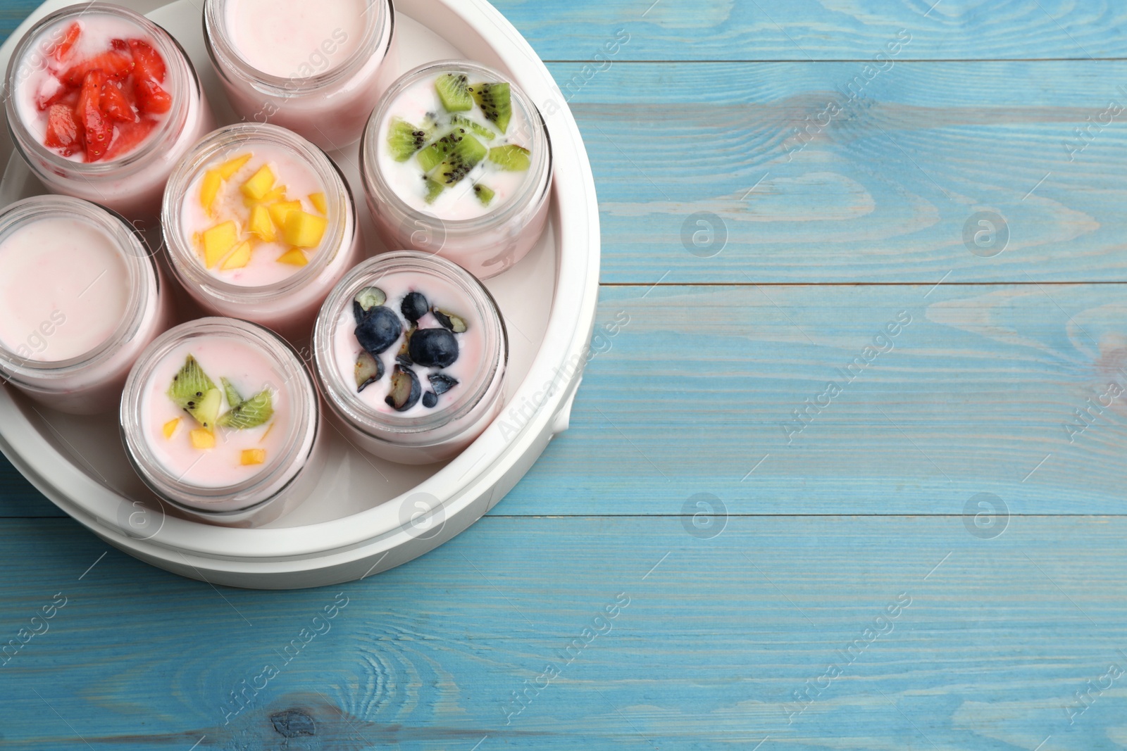 Photo of Modern yogurt maker with full jars and different fruits on light blue wooden table, top view. Space for text