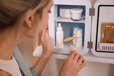 Photo of Woman with face roller taking cosmetic product from mini fridge indoors, closeup