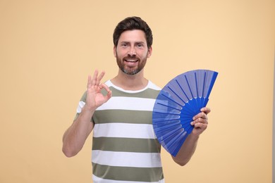 Photo of Happy man holding hand fan and showing ok gesture on beige background