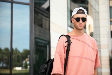 Photo of Handsome young man with stylish sunglasses and backpack near building outdoors, space for text