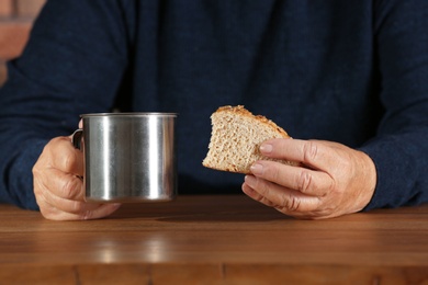 Poor elderly woman with piece of bread and metal mug at table, focus on hands