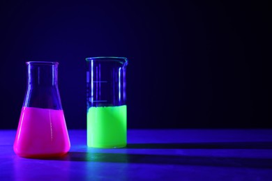 Laboratory glassware with luminous liquids on table against dark background, space for text