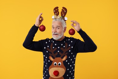 Photo of Senior man in Christmas sweater and reindeer headband holding festive baubles on orange background