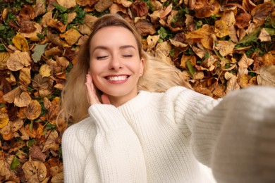Smiling woman lying among autumn leaves and taking selfie outdoors, top view