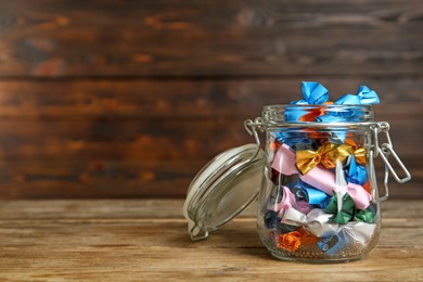 Photo of Glass jar with candies in colorful wrappers on wooden table. Space for text