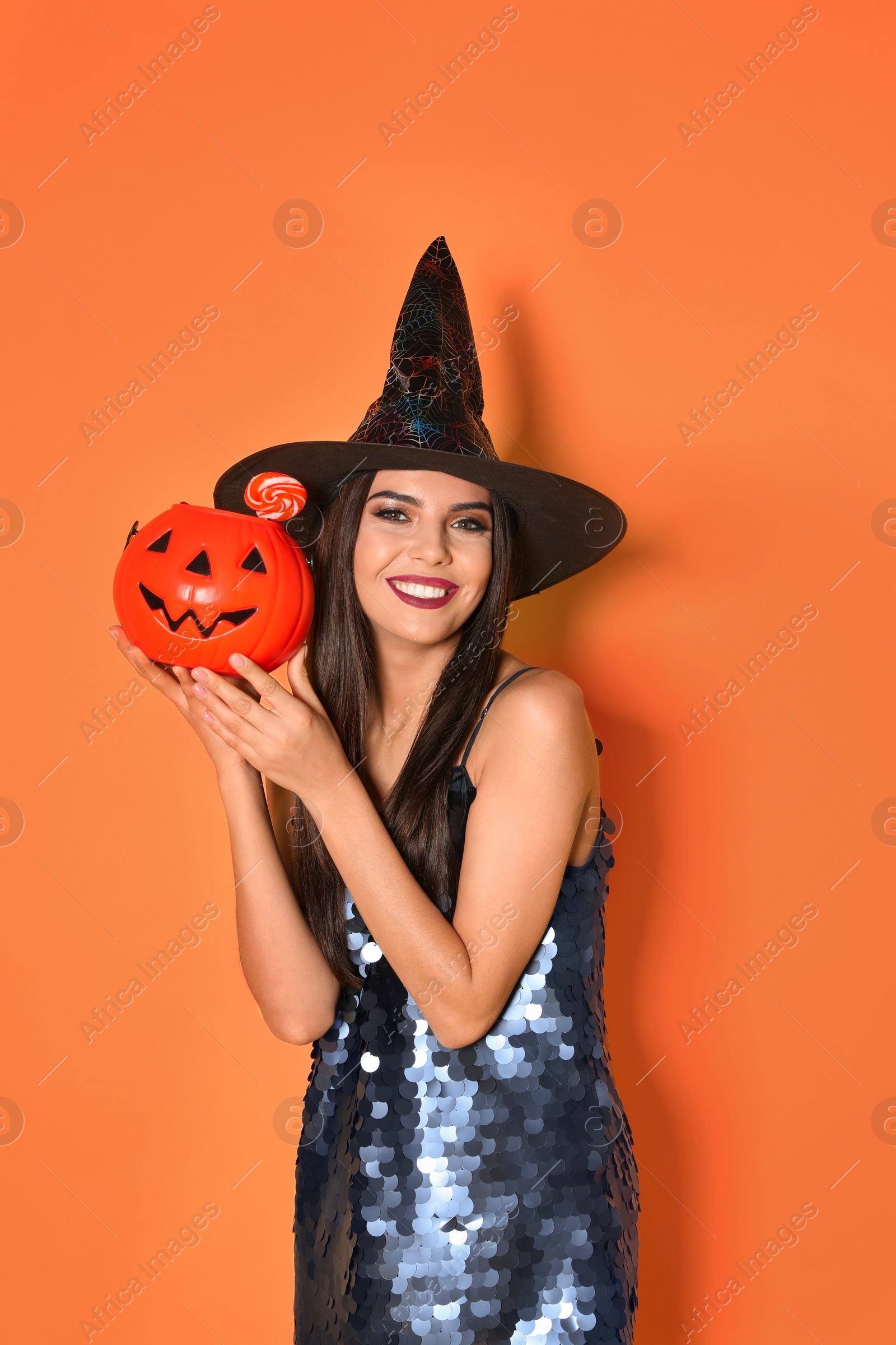 Photo of Beautiful woman wearing witch costume with Jack O'Lantern candy container for Halloween party on yellow background
