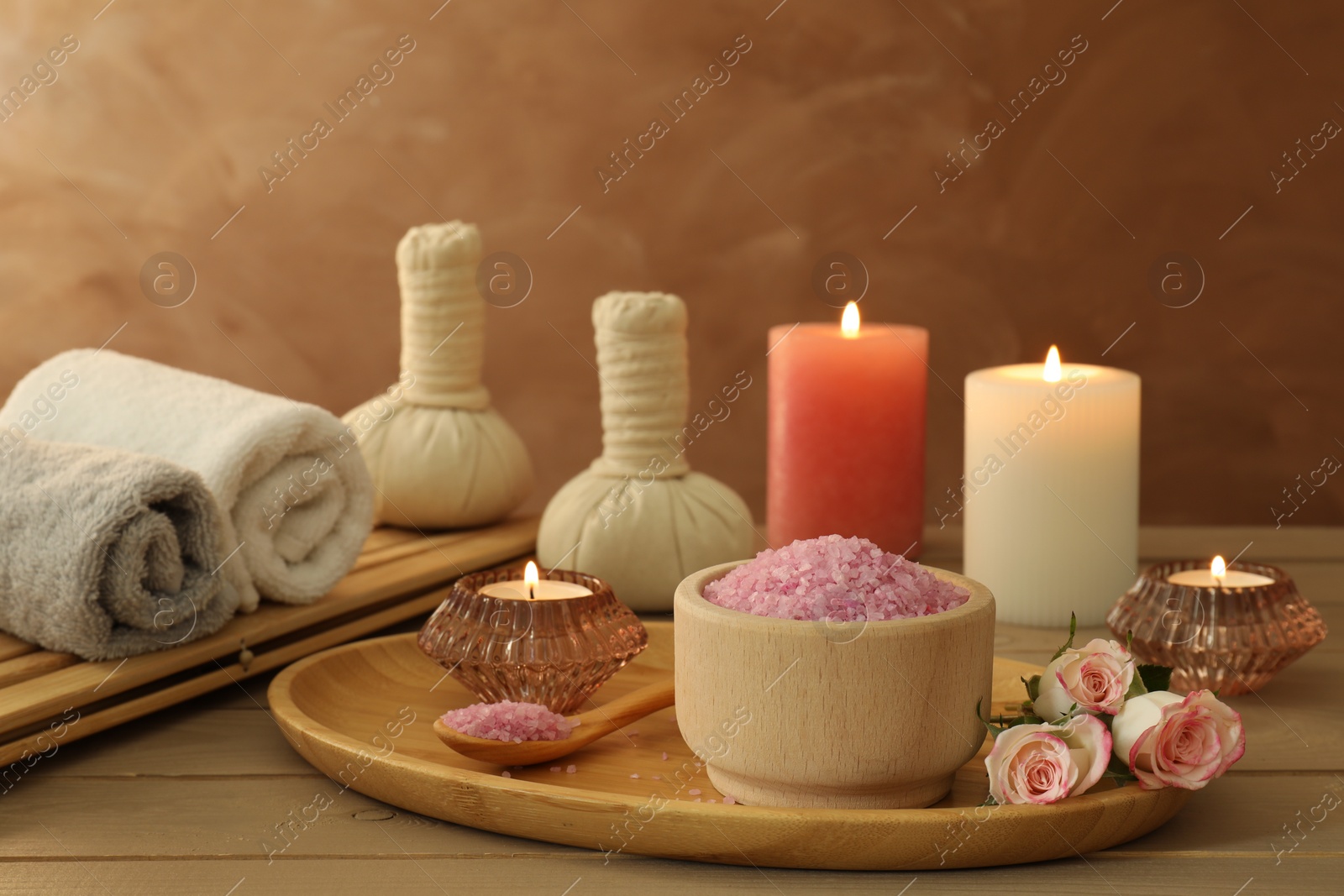 Photo of Bowl of pink sea salt, roses, burning candles, herbal massage bags and towels on wooden table
