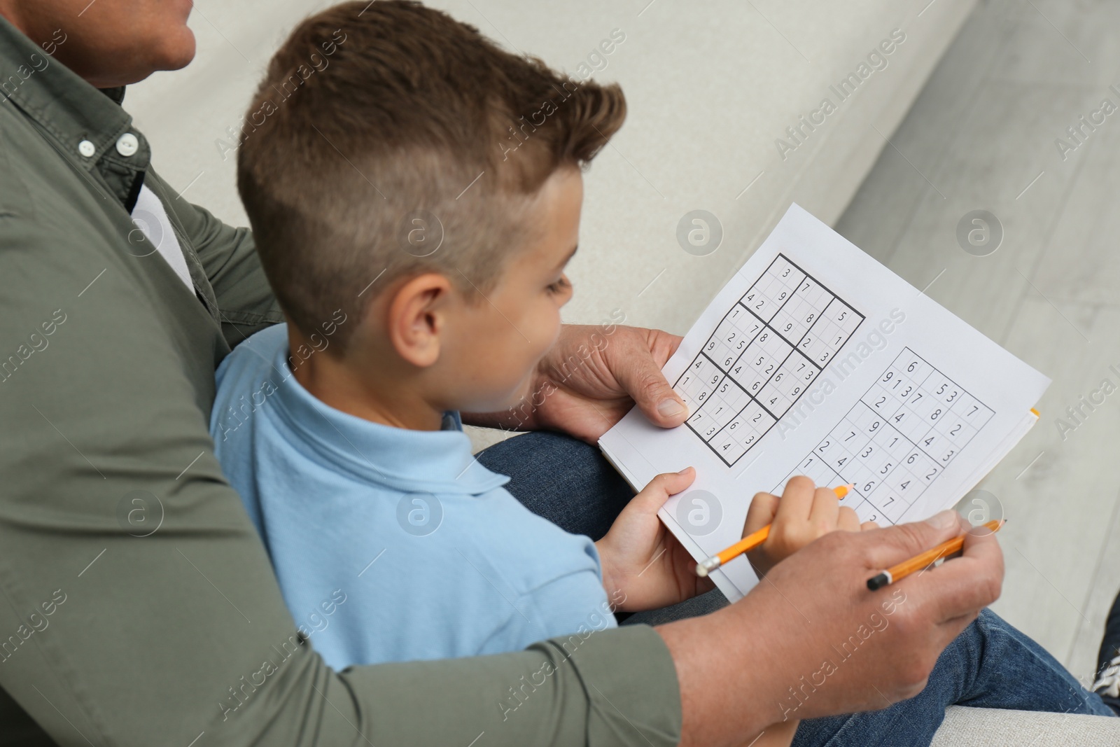 Photo of Little boy with his grandfather solving sudoku puzzle on sofa at home, closeup