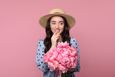 Beautiful young woman in straw hat with bouquet of peonies on pink background