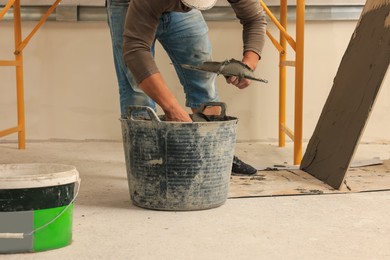 Worker applying adhesive mix on spatula near tile indoors, closeup