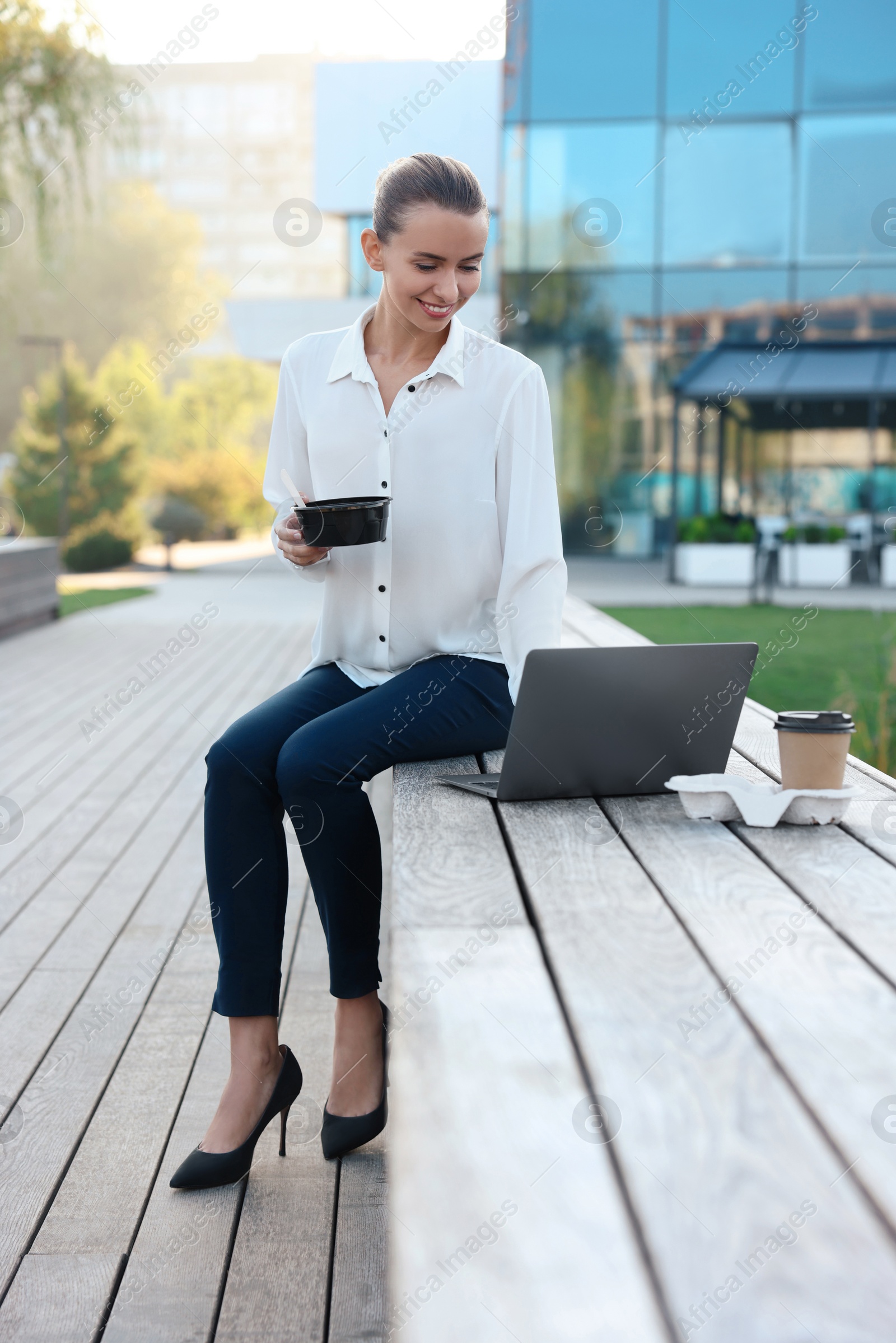 Photo of Smiling businesswoman holding lunch box and working with laptop outdoors