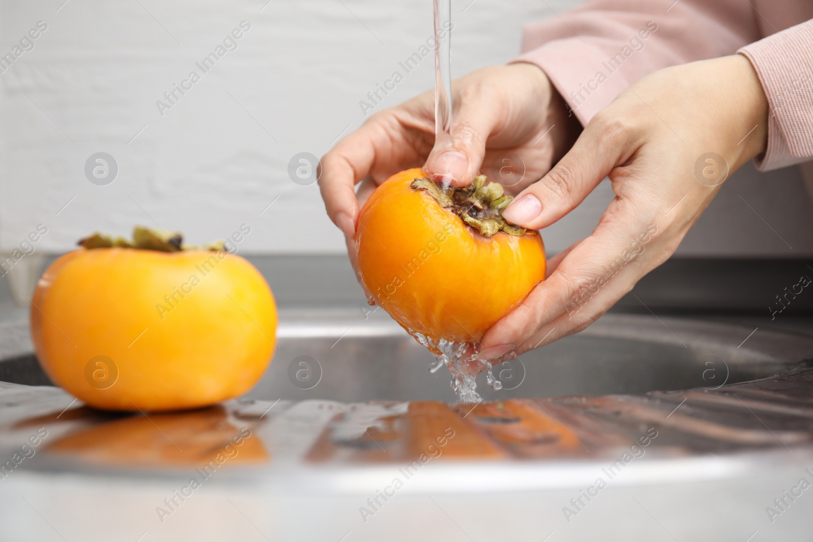 Photo of Woman washing delicious ripe juicy persimmons under tap water indoors, closeup