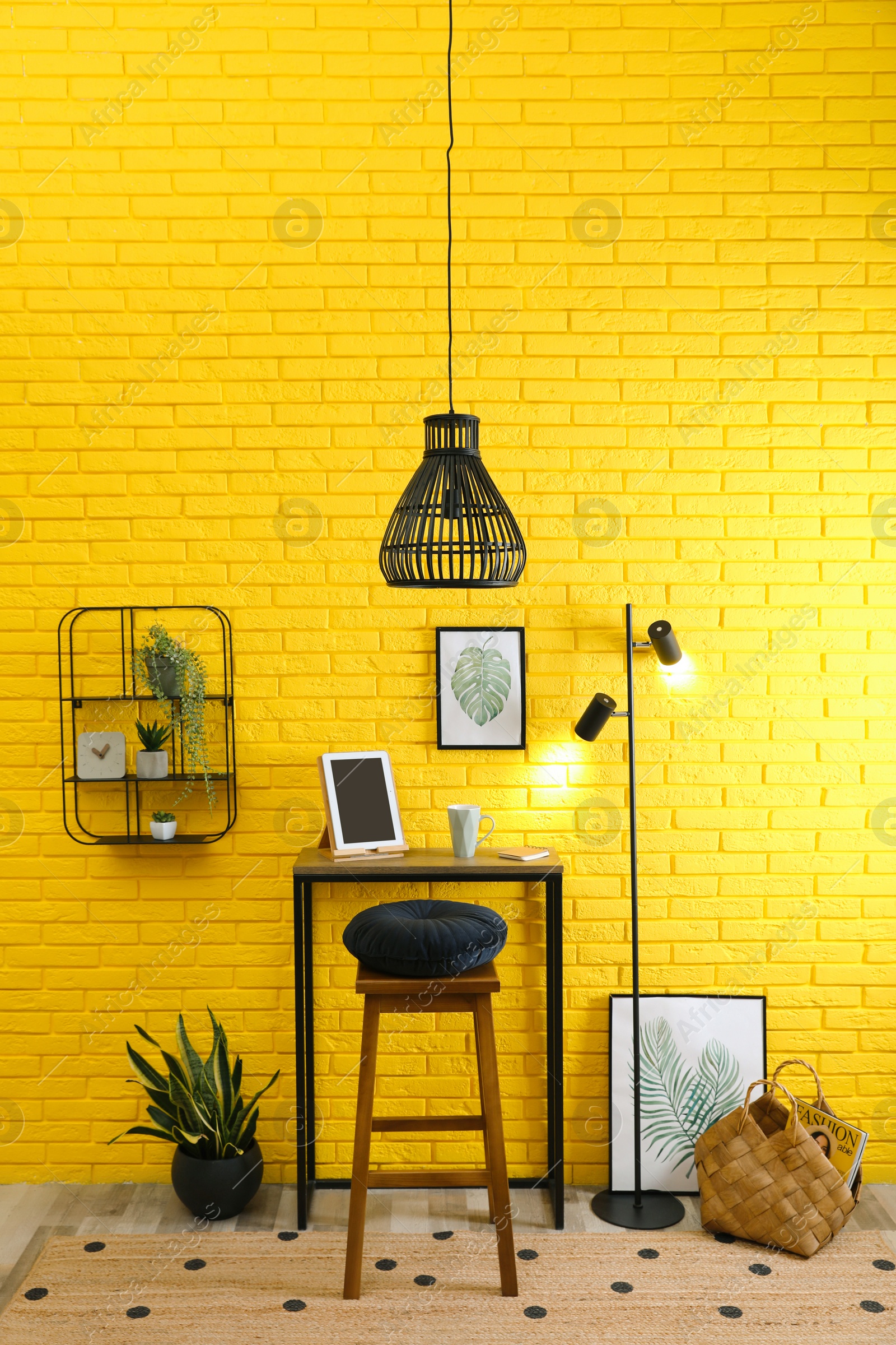 Photo of High wooden table with tablet and stool as stand up workplace near yellow brick wall. Stylish interior