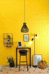 High wooden table with tablet and stool as stand up workplace near yellow brick wall. Stylish interior