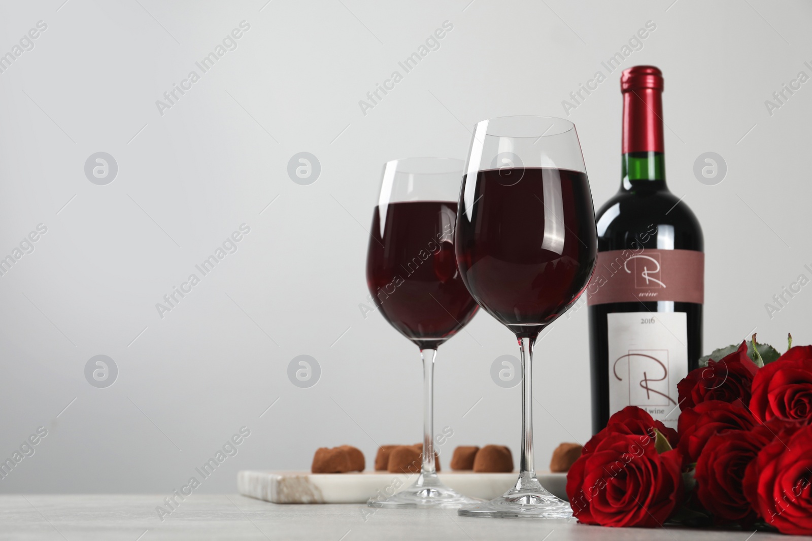 Photo of Bottle with glasses of red wine, beautiful roses and chocolate candies on white table. Space for text