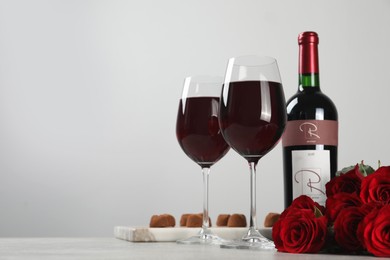 Bottle with glasses of red wine, beautiful roses and chocolate candies on white table. Space for text