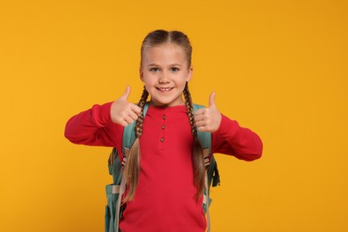 Photo of Happy schoolgirl with backpack showing thumbs up gesture on orange background